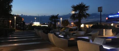 An outdoor seating area with modern chairs and tables, a palm tree, and a building with a sign in the background.