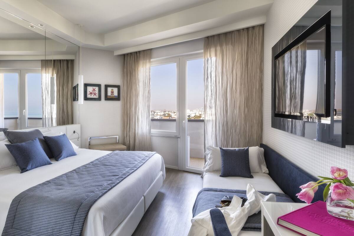 A modern hotel room with a large bed, a seating area, a balcony with a view, and a flat-screen TV.