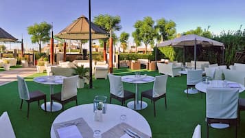 Een terras met ronde tafels en stoelen, een pergola en groen.