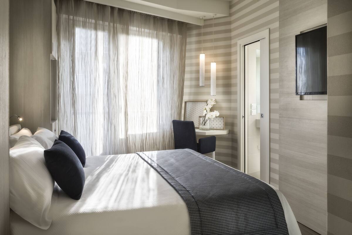 A modern hotel room with a large bed, a desk, and a large window with sheer curtains.