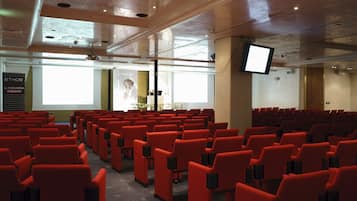 Una sala conferenze con file di sedie rosse, un grande schermo e un proiettore.