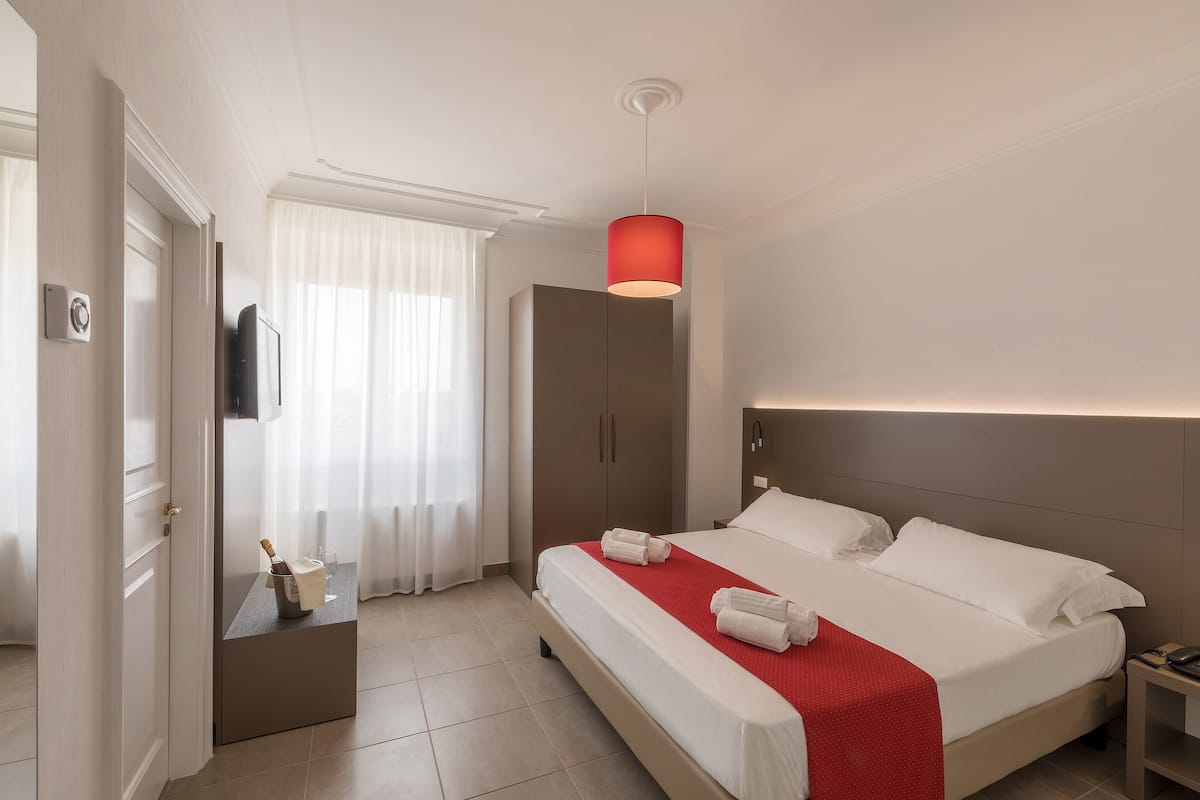 A modern hotel room with a large bed, a bedside table with a bottle and glasses, a wardrobe, and a red pendant light.