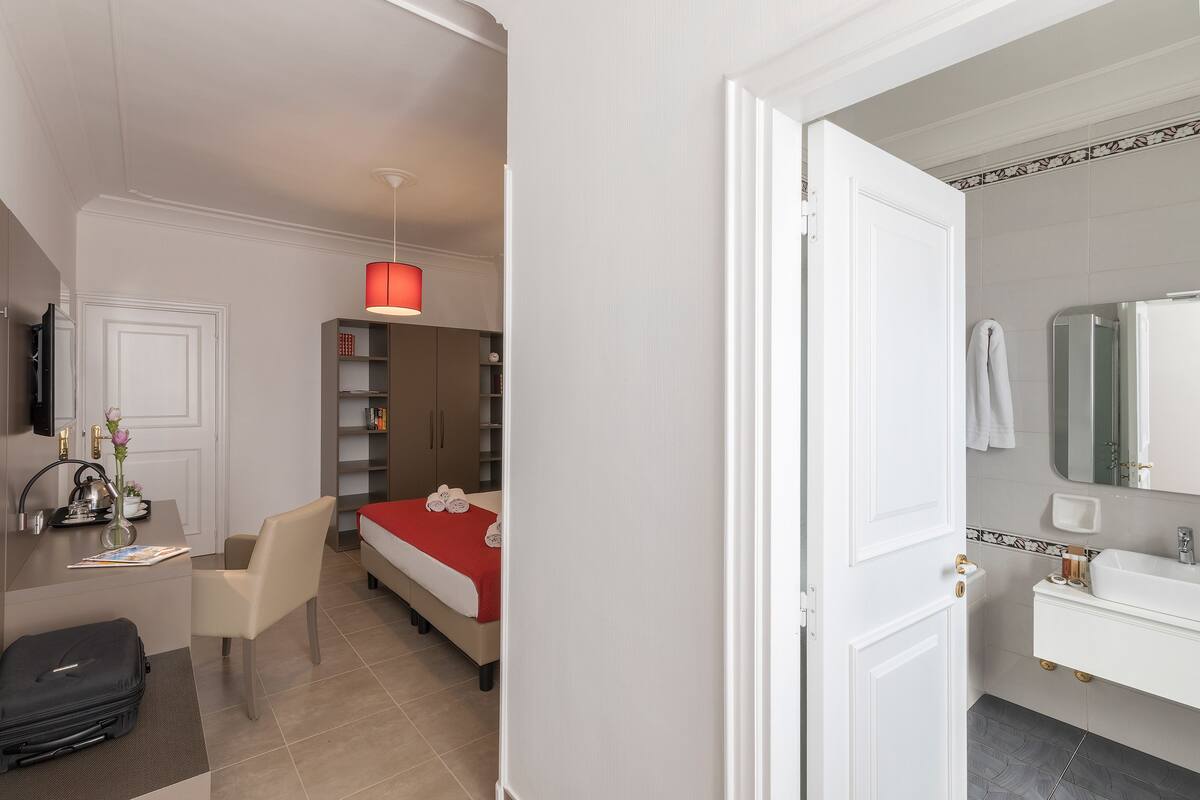 A hotel room with a bed, a desk, a chair, a suitcase, a red hanging lamp, and a bathroom with a sink and mirror.