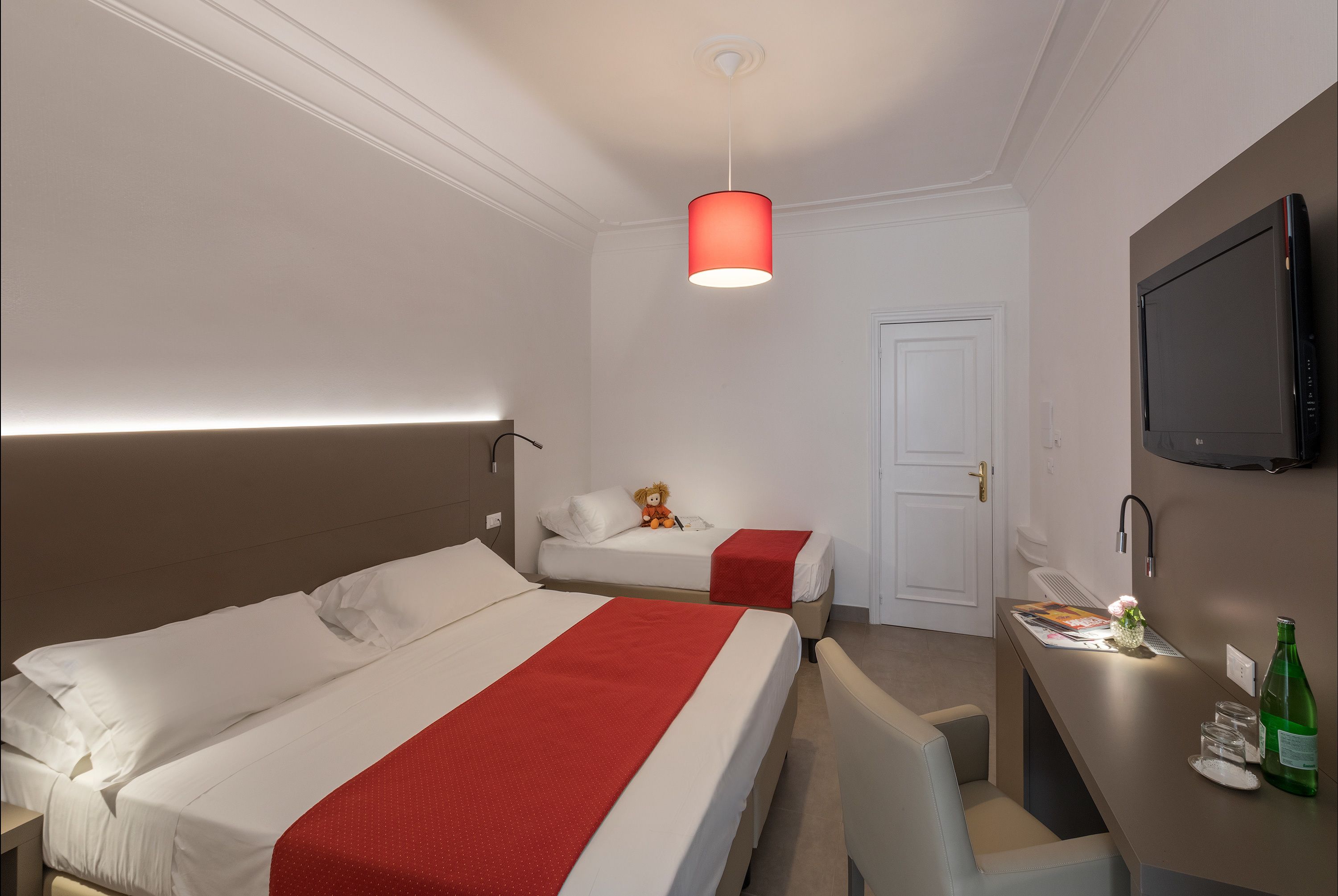Une chambre d’hôtel avec deux lits, un bureau, une chaise, une télévision et une lampe suspendue rouge.