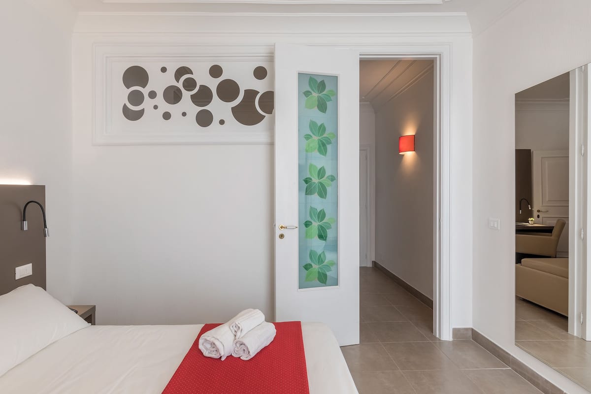 A hotel room with a neatly made bed, a red accent pillow, a wall with abstract art, and a door leading to a bathroom with a towel rack.