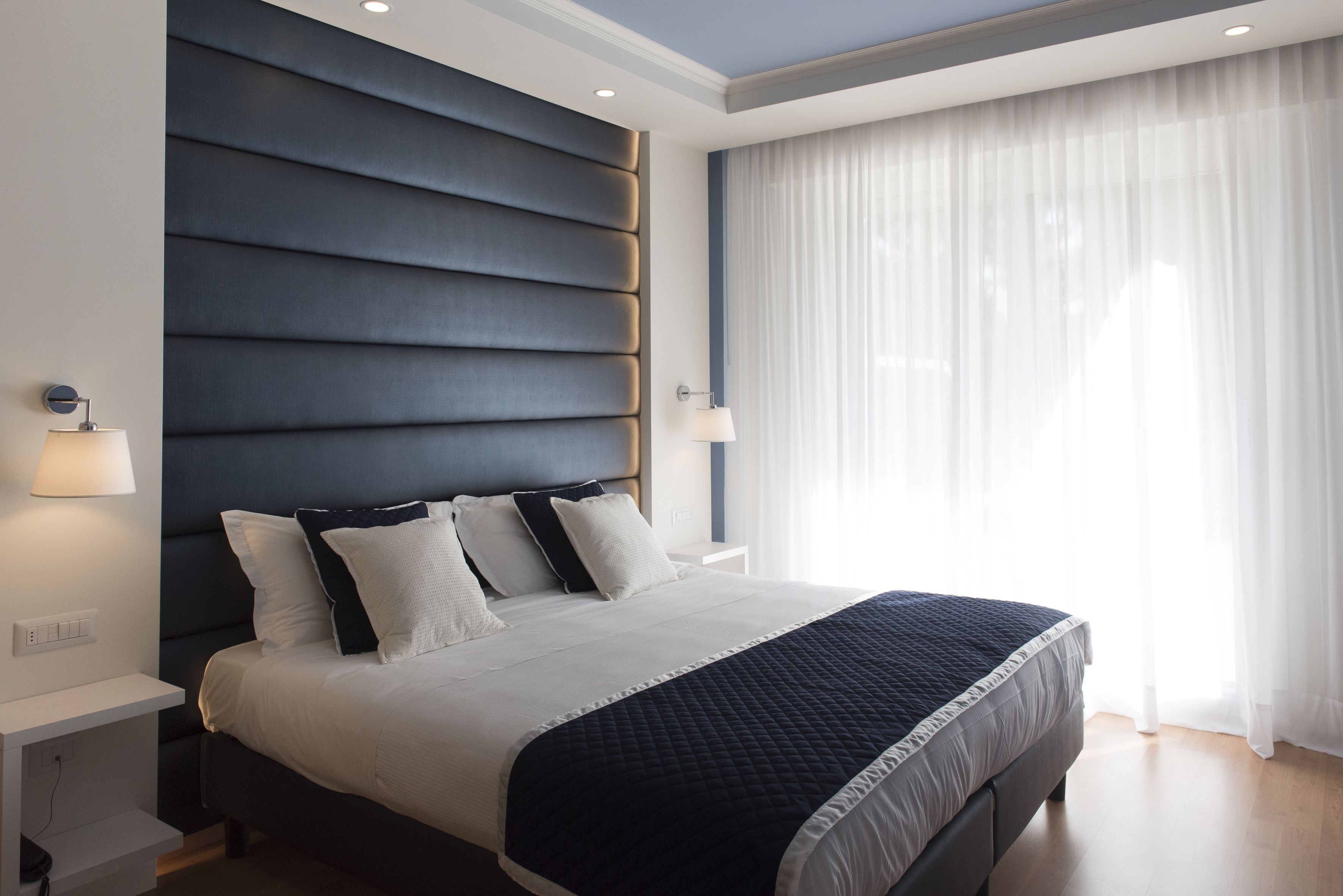 Un dormitorio moderno con una cama grande, un cabecero oscuro, lámparas de noche y cortinas translúcidas.
