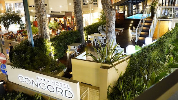 A hotel lobby with a pool area, outdoor seating, and a sign for the Concord Hotel. - Hotel Concord (Riccione)