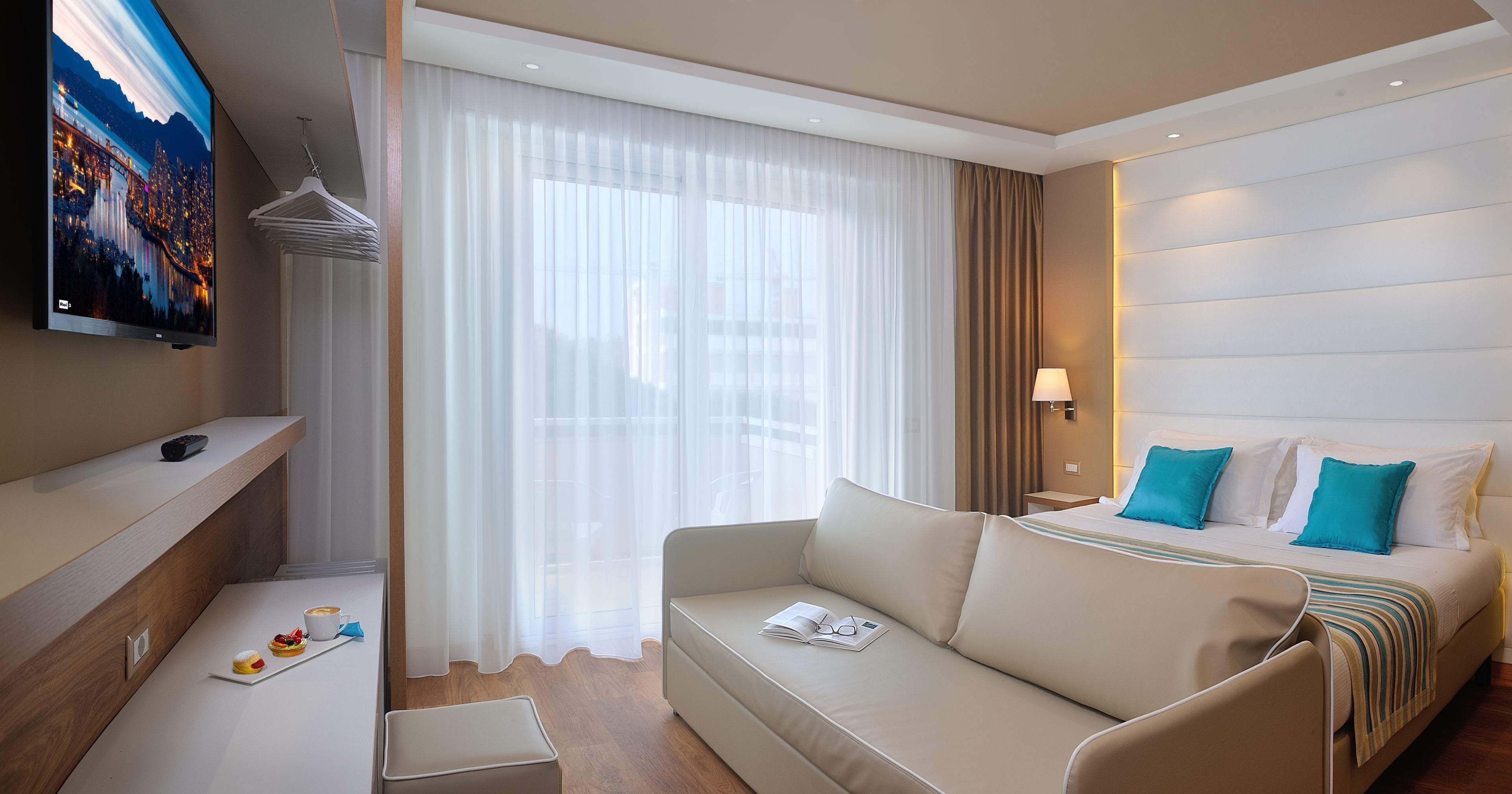 A modern hotel room with a sofa, a bed with blue pillows, a TV, a coffee table, and a window with sheer curtains.