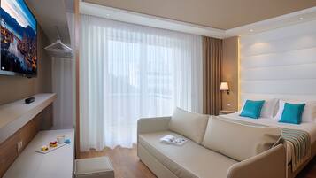 A modern hotel room with a sofa, a bed with blue pillows, a TV, a coffee table, and a window with sheer curtains.