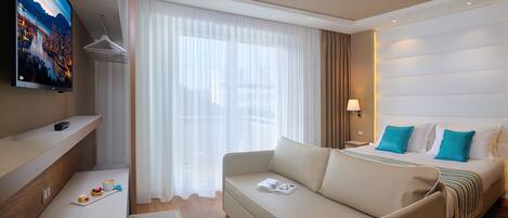 A modern hotel room with a sofa, a bed with blue pillows, a TV, a coffee table, and a window with sheer curtains.