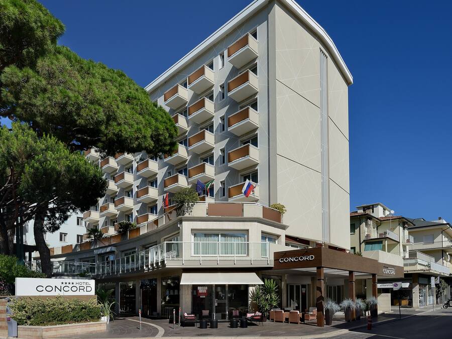 Un hotel de varios pisos con balcones, un cartel que dice 'CONCORD HOTEL' y zonas de estar al aire libre.
