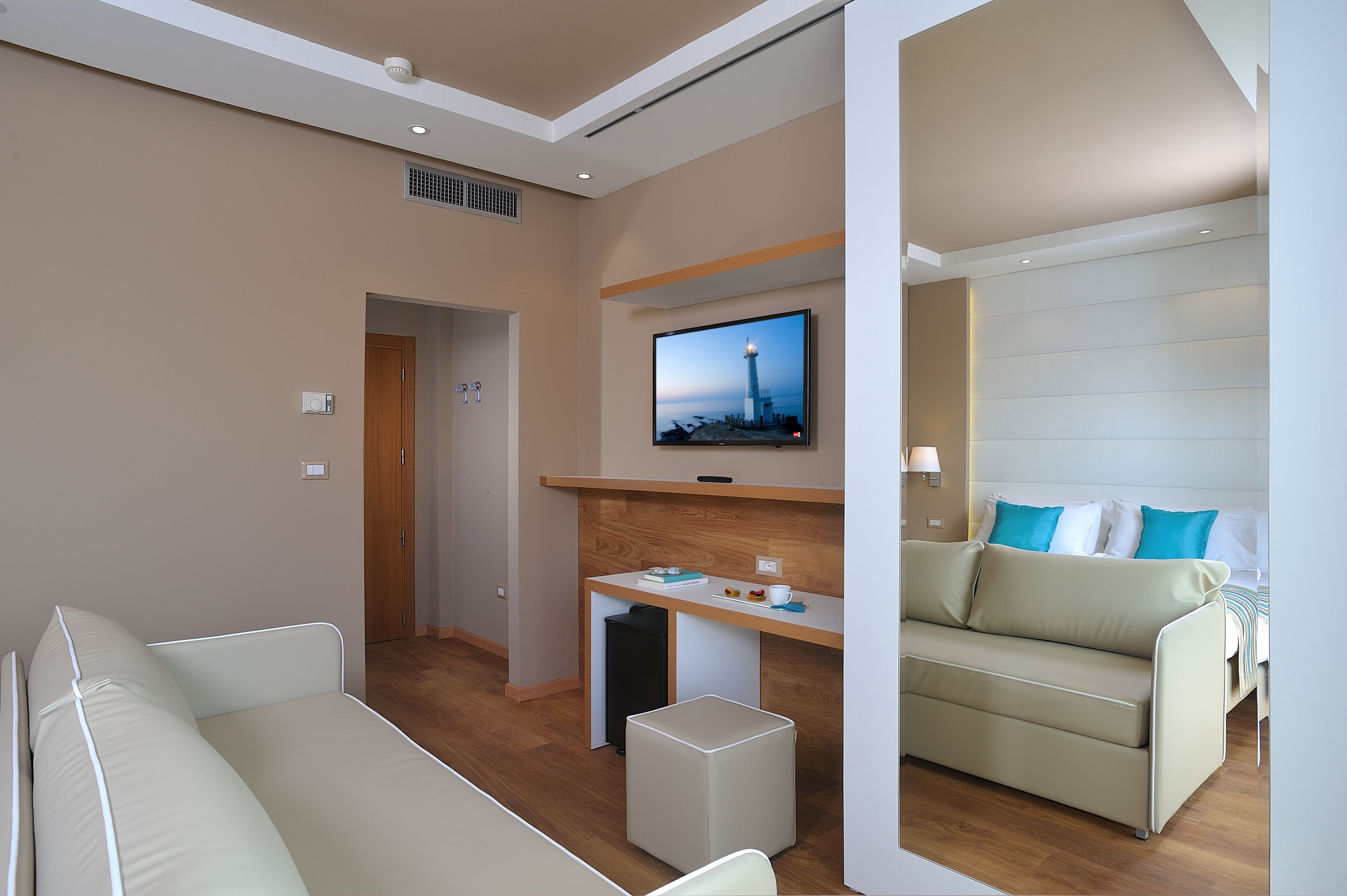 A modern hotel room with a bed, a sofa, a desk with a TV, and a small kitchenette.