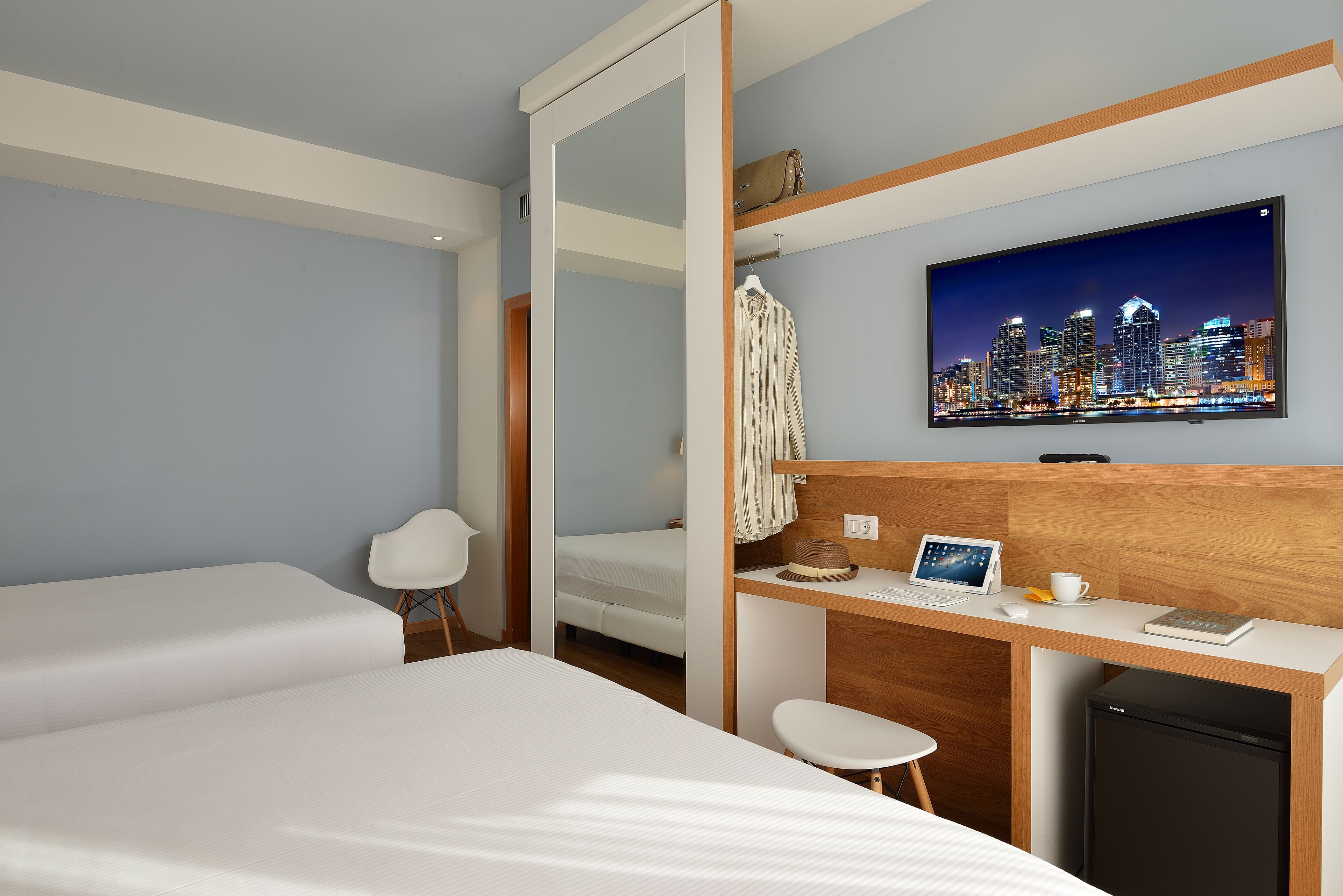 Habitación de hotel con cama, escritorio con computadora portátil, refrigerador pequeño y cuadro de un paisaje urbano en la pared.