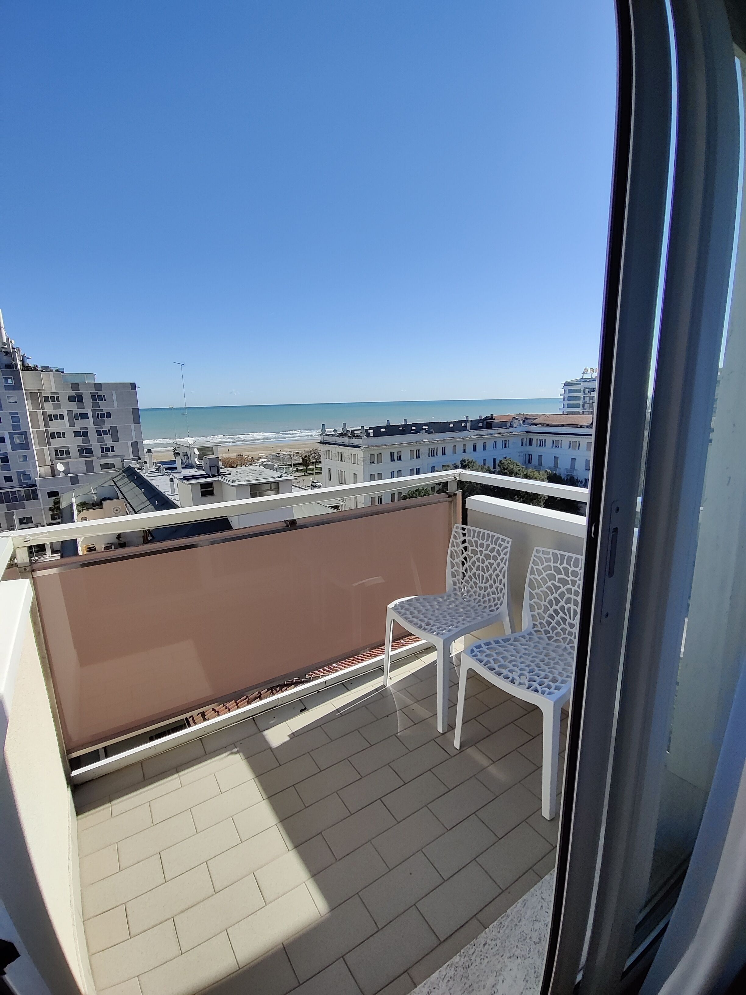 A balcony with two white chairs, a view of the beach, and buildings in the background.