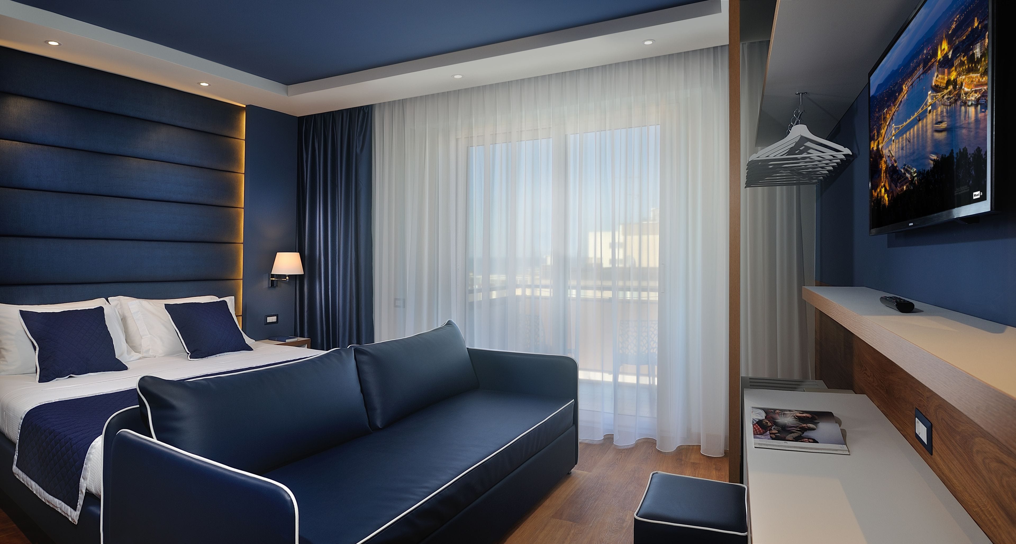 Una camera d'albergo moderna con un letto grande, un divano, una TV a parete e un comodino in legno.