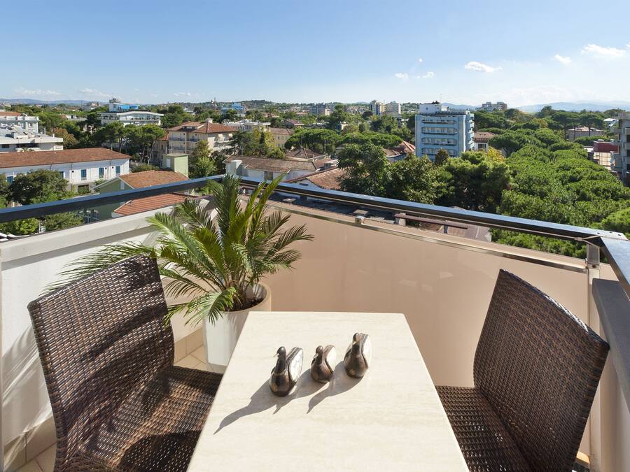 Un balcone con sedie in vimini e un tavolo, affacciato su un panorama urbano con edifici e vegetazione.