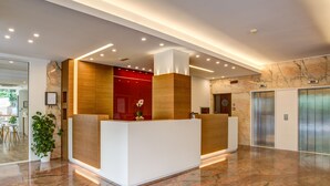 A modern reception area with a marble floor, wooden reception desk, and a red accent wall. - Hotel Concord (Riccione)