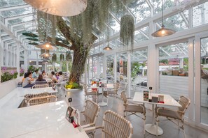 A spacious indoor dining area with high ceilings and large windows, featuring wicker furniture and hanging plants. - Sino House Phuket Hotel (Phuket)