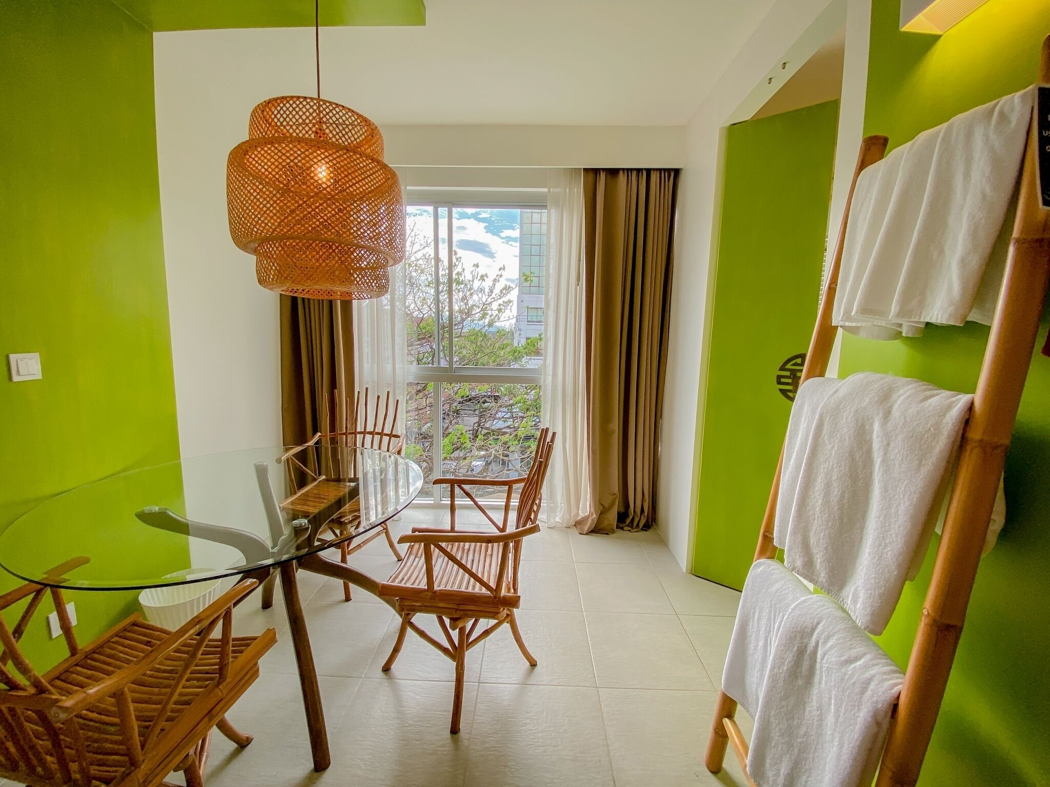 Une pièce moderne avec une table en verre, des chaises en bois, un luminaire en fibres tressées et une vue dégagée sur l’extérieur grâce à une grande fenêtre.