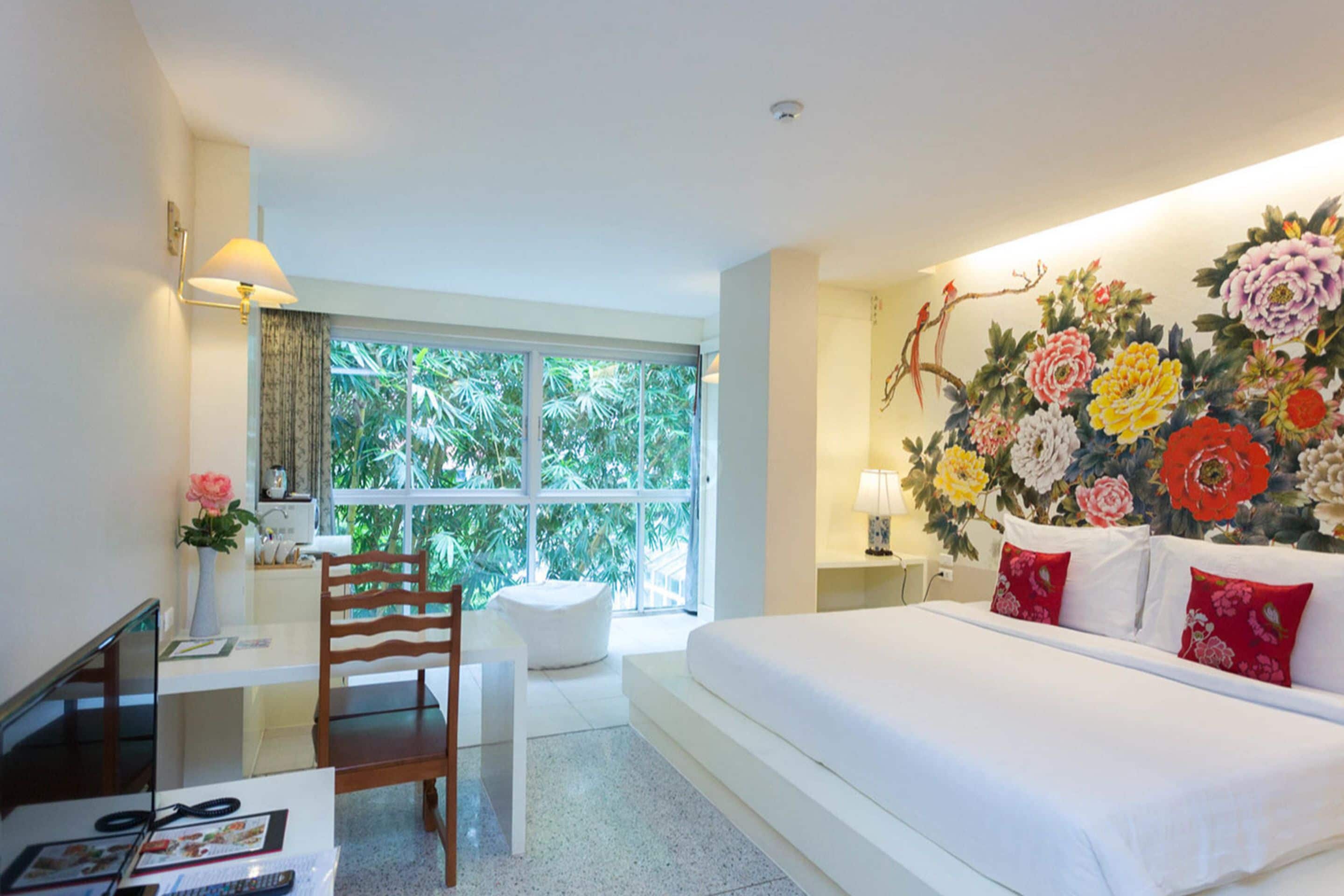 A hotel room with a large bed, a desk with a chair, a window with a view of greenery, and a wall decorated with flowers.