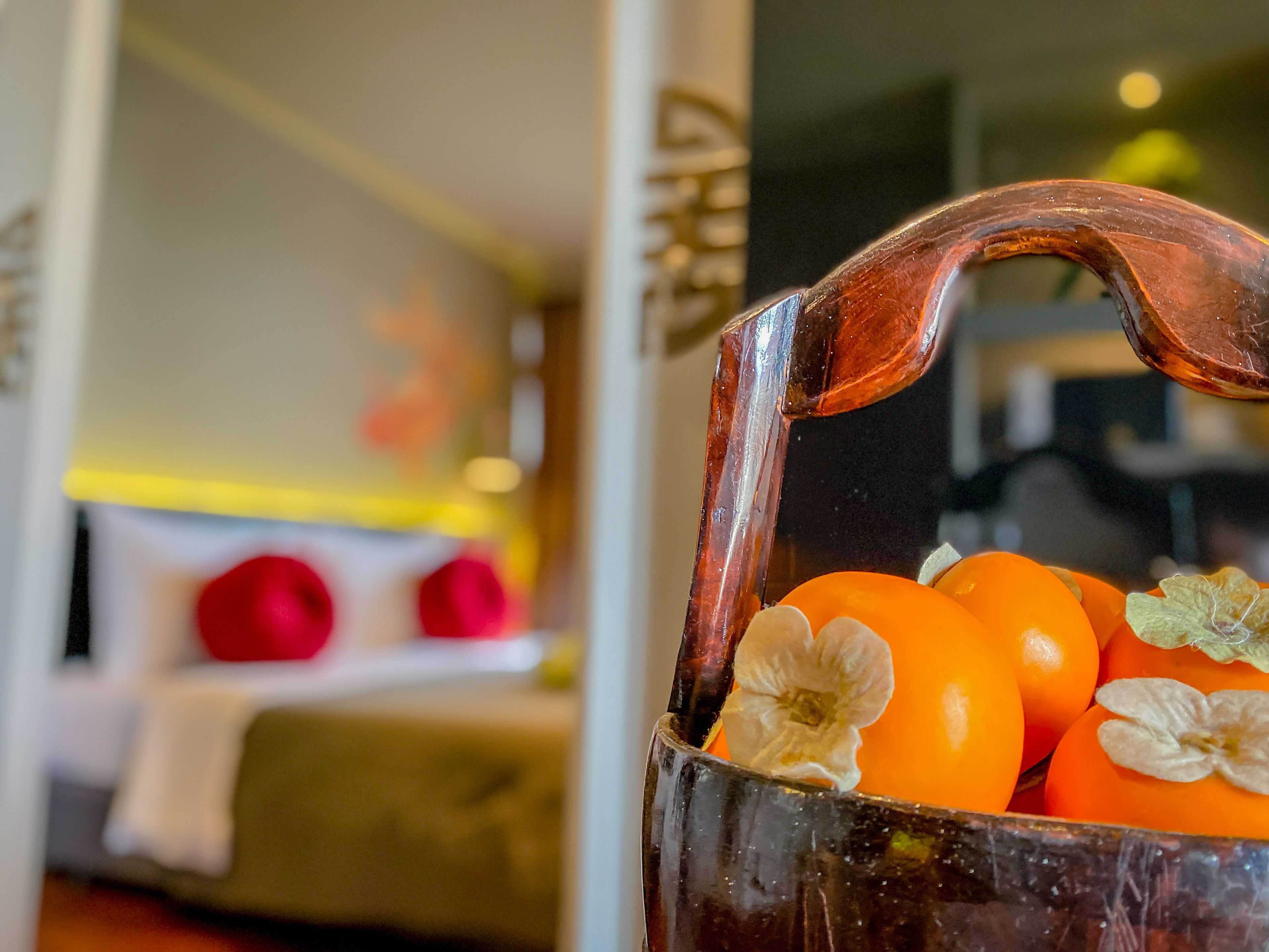 A close-up of a copper-colored vase with orange fruits and white flowers, placed in a room with a bed and pillows in the background.