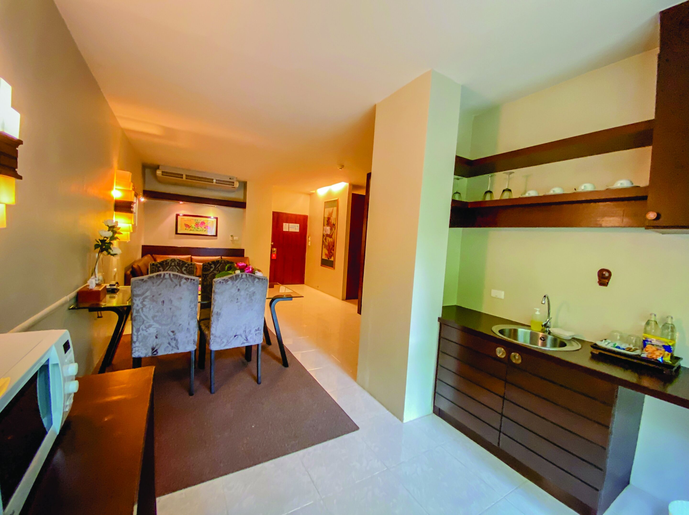 Una habitación de hotel con comedor, una cocina que incluye un lavamanos y armarios, y una puerta que da a otra habitación.