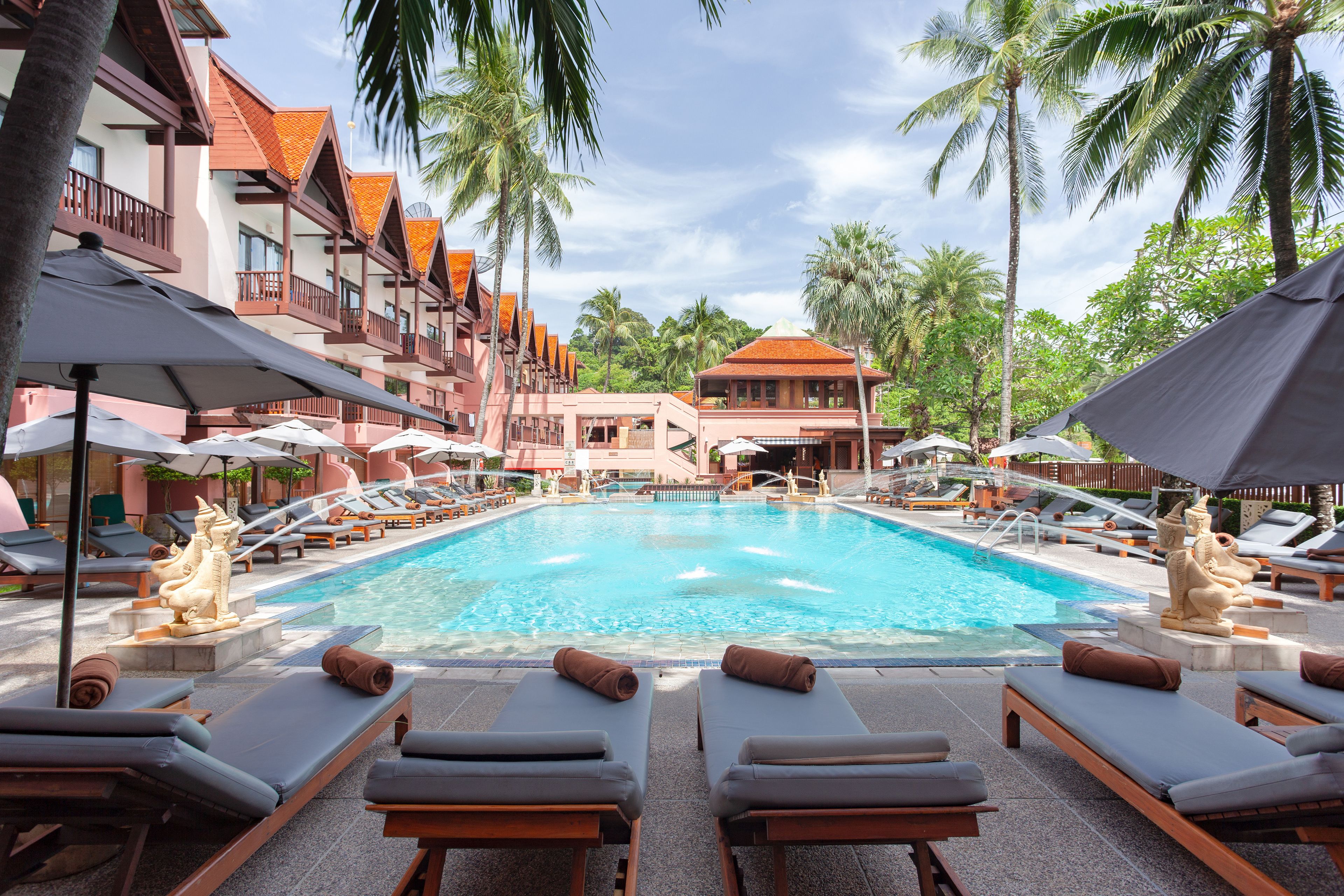 A pool area with lounge chairs and umbrellas, surrounded by palm trees and a building with a red roof.
