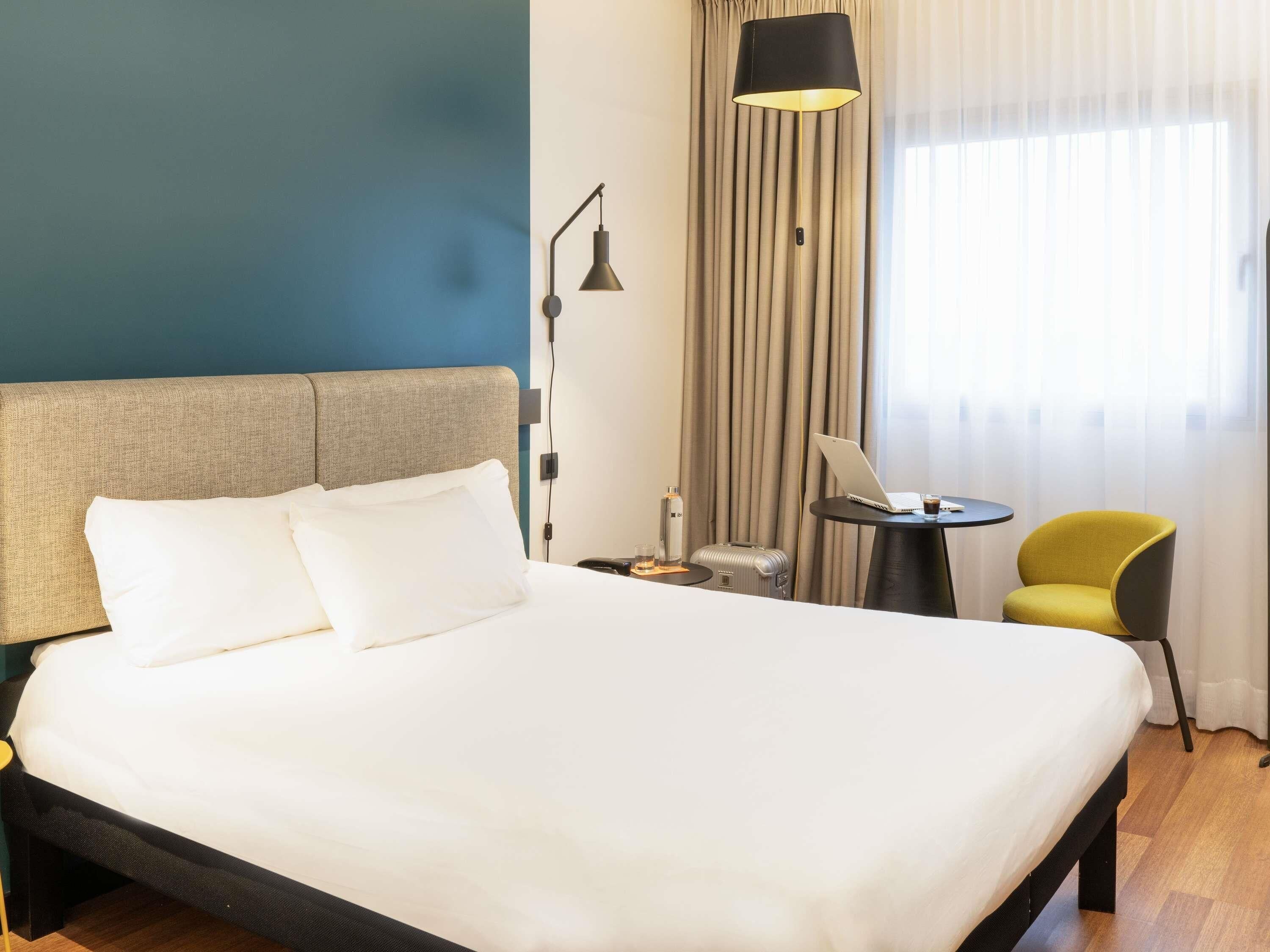 A hotel room with a bed, a desk, a chair, a lamp, and a window with curtains.