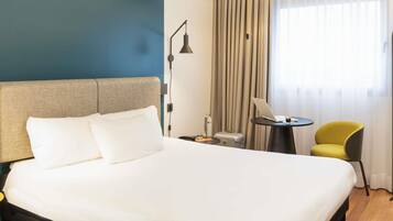 A hotel room with a bed, a desk, a chair, a lamp, and a window with curtains.