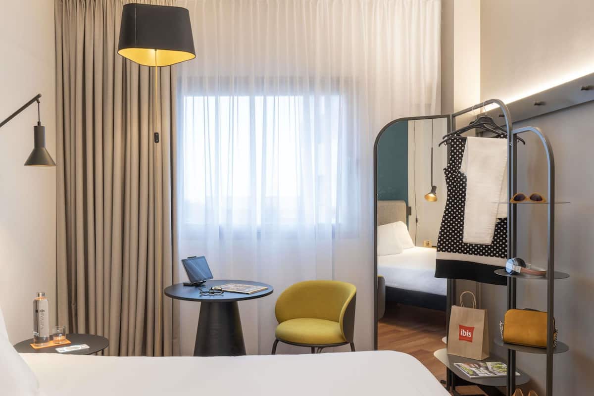 A hotel room with a bed, a desk, a chair, a hanging rack with clothes and shoes, and a window with curtains.