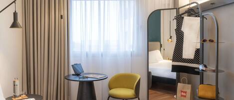 Quarto de hotel com cama, escrivaninha, cadeira, cabideiro com roupas e sapatos, e janela com cortinas.