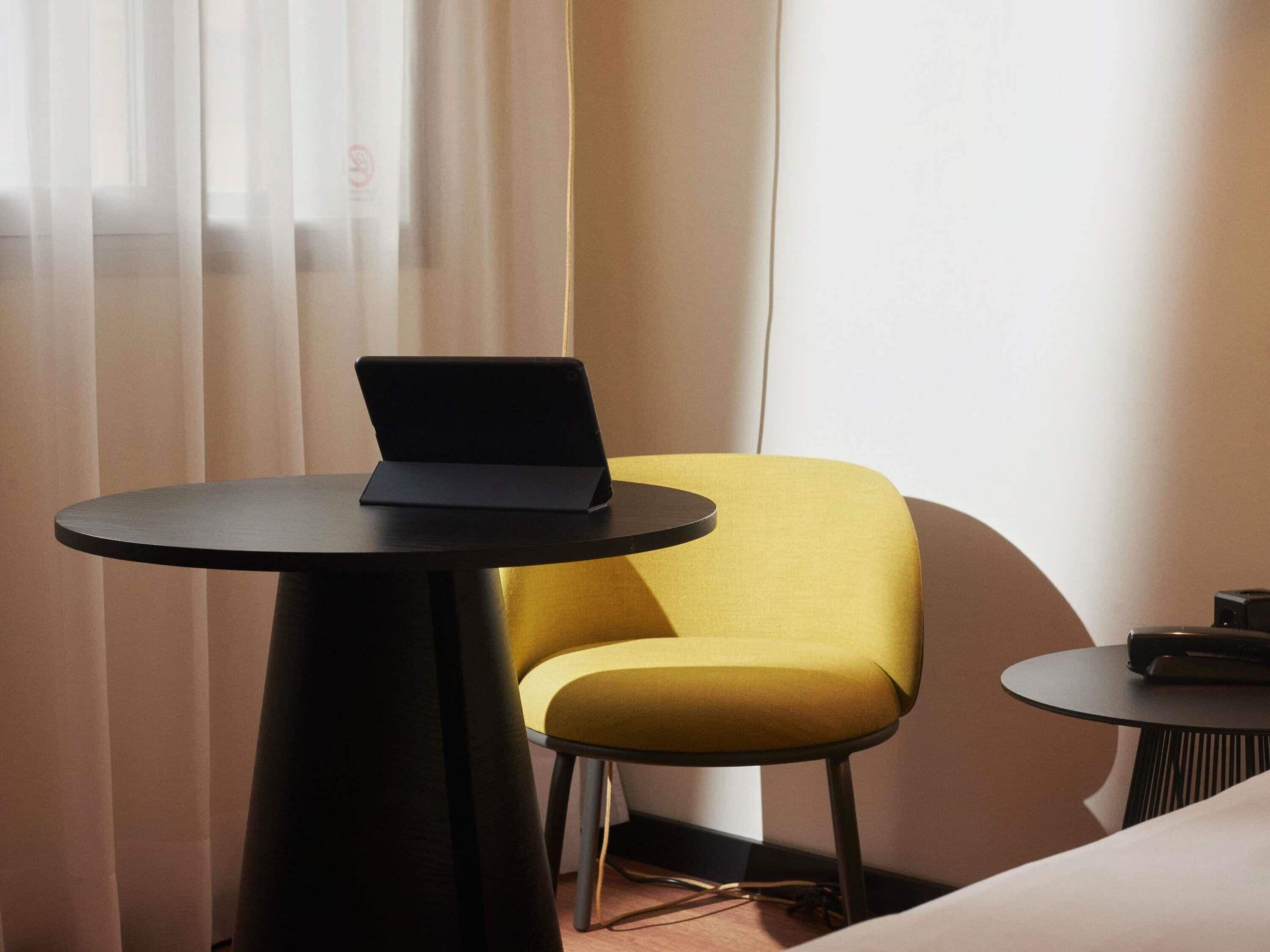 A hotel room with a bed, a small round table with a laptop, a yellow armchair, and a second table with a remote control.