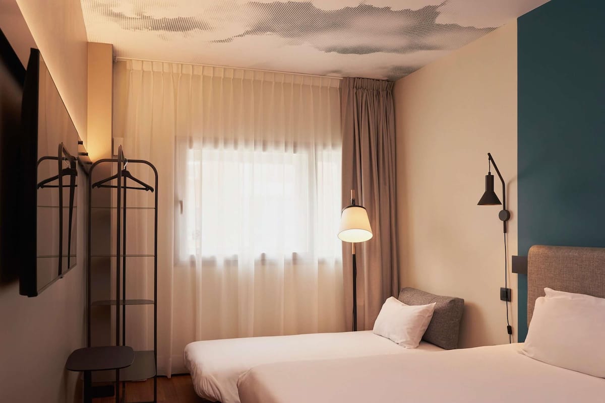 A hotel room with a bed, a desk, a chair, a wardrobe, a window with curtains, and a lamp.
