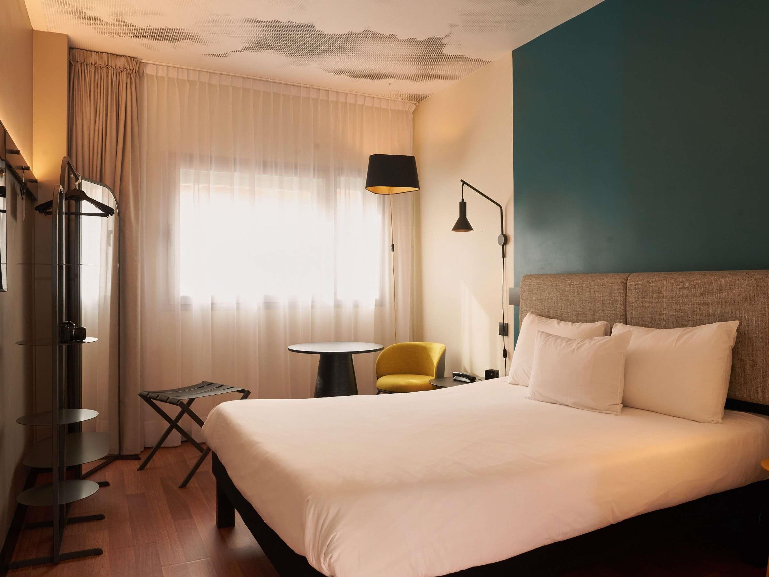 A hotel room with a bed, a small table, a chair, a lamp, and a coat rack.
