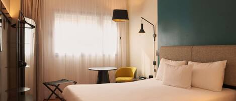 A hotel room with a bed, a small table, a chair, a lamp, and a coat rack.