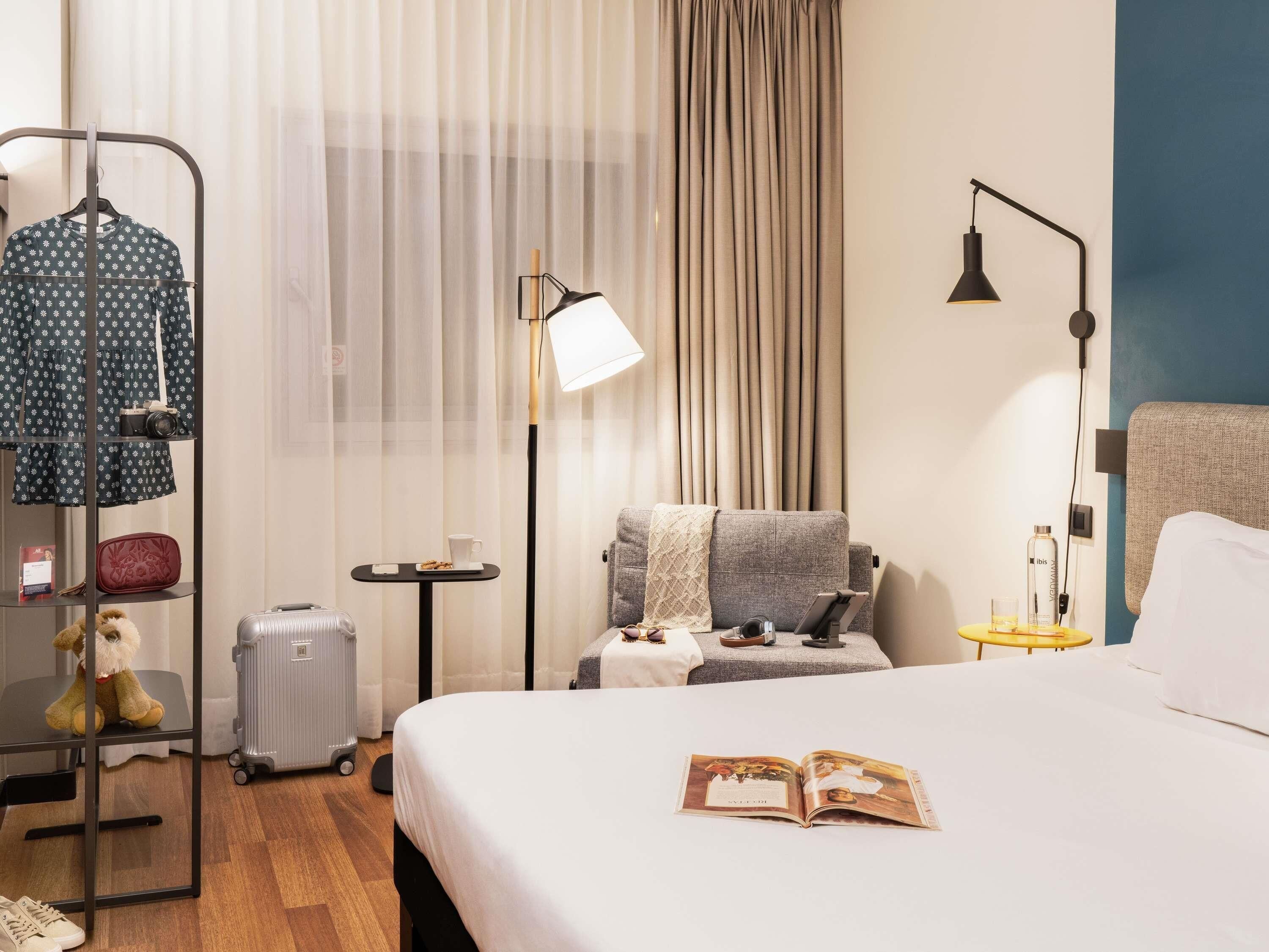 Une chambre d’hôtel avec un lit, un coin lecture avec une lampe, une petite table avec une tasse et un livre, une armoire contenant des vêtements, un ours en peluche et une valise.