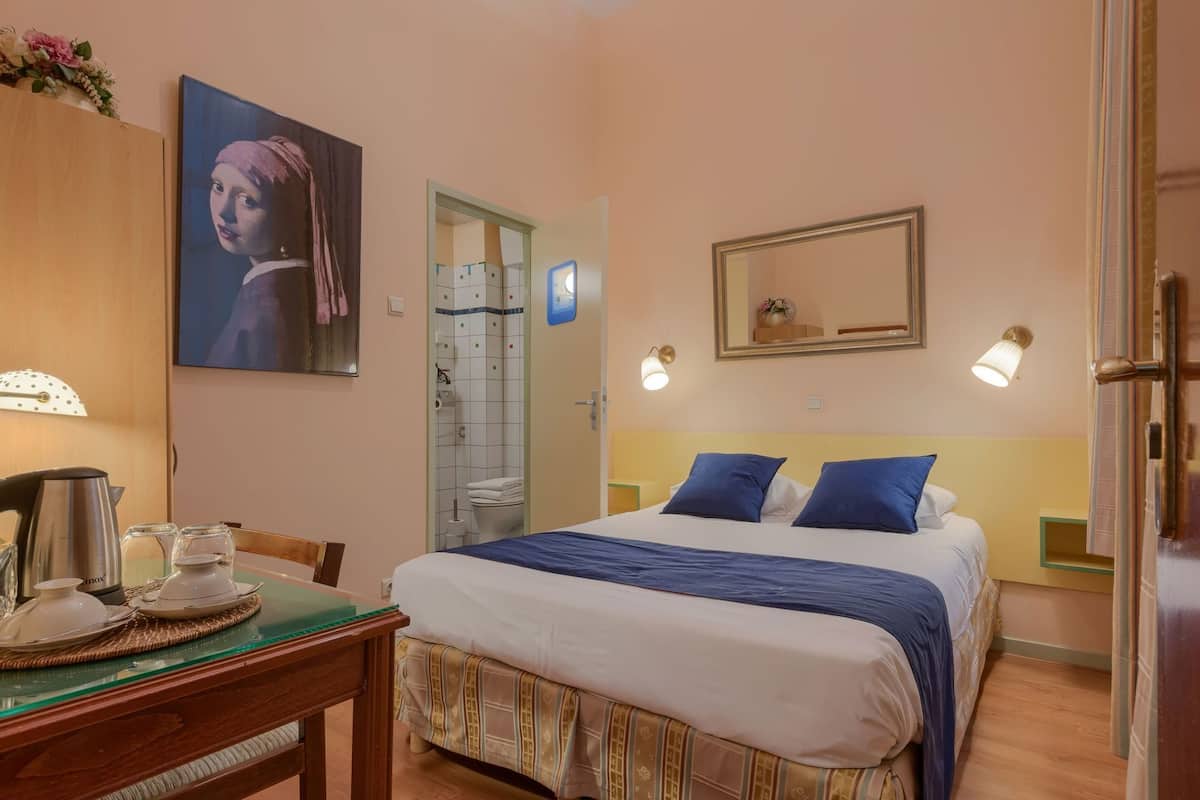 A hotel room with a bed, bedside tables, a desk with a kettle, a mirror, and a painting.