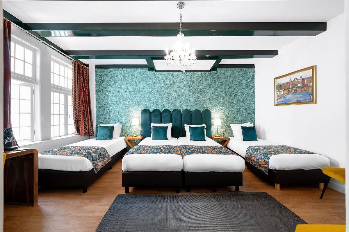 A hotel room with a large bed, two smaller beds, a chandelier, a painting, and a view of the city.