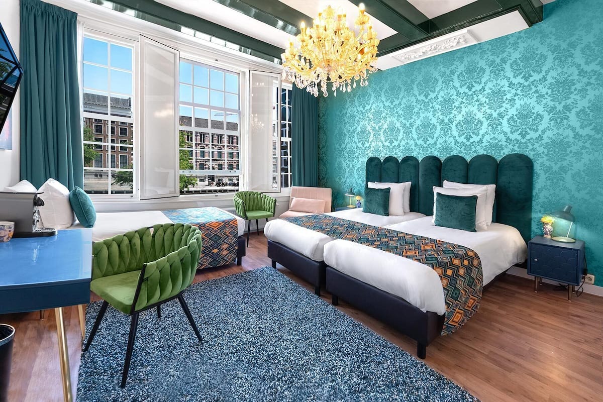 A modern bedroom with a large bed, a desk, and a chair. There is a view of buildings outside the window.