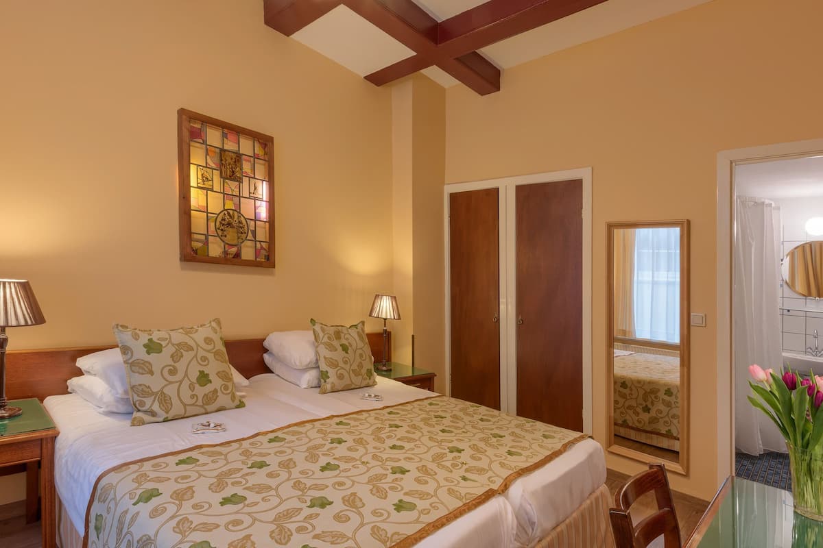 A hotel room with a bed, two bedside lamps, a stained glass window, a wooden door, a mirror, and a vase with flowers.