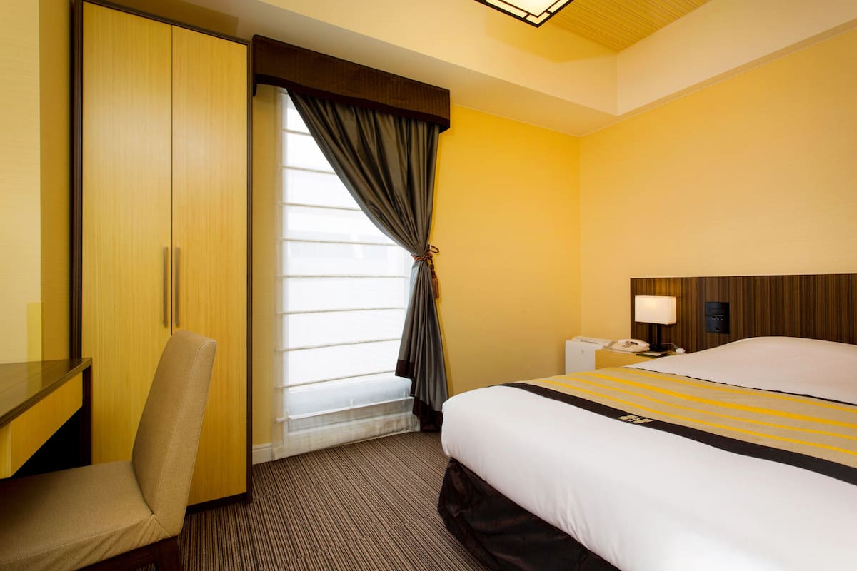 A hotel room with a bed, a desk, a chair, a window with curtains, a nightstand, and a lamp.