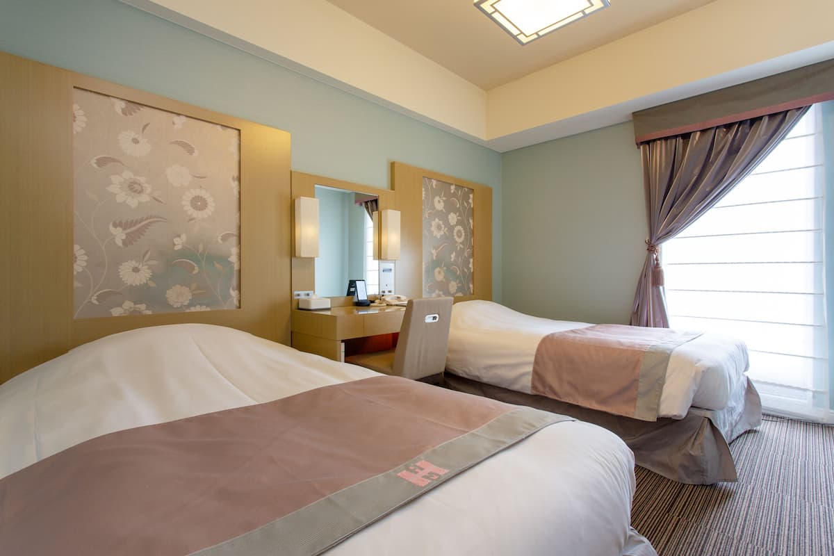 A hotel room with two beds, a desk, a chair, and a large window with curtains.