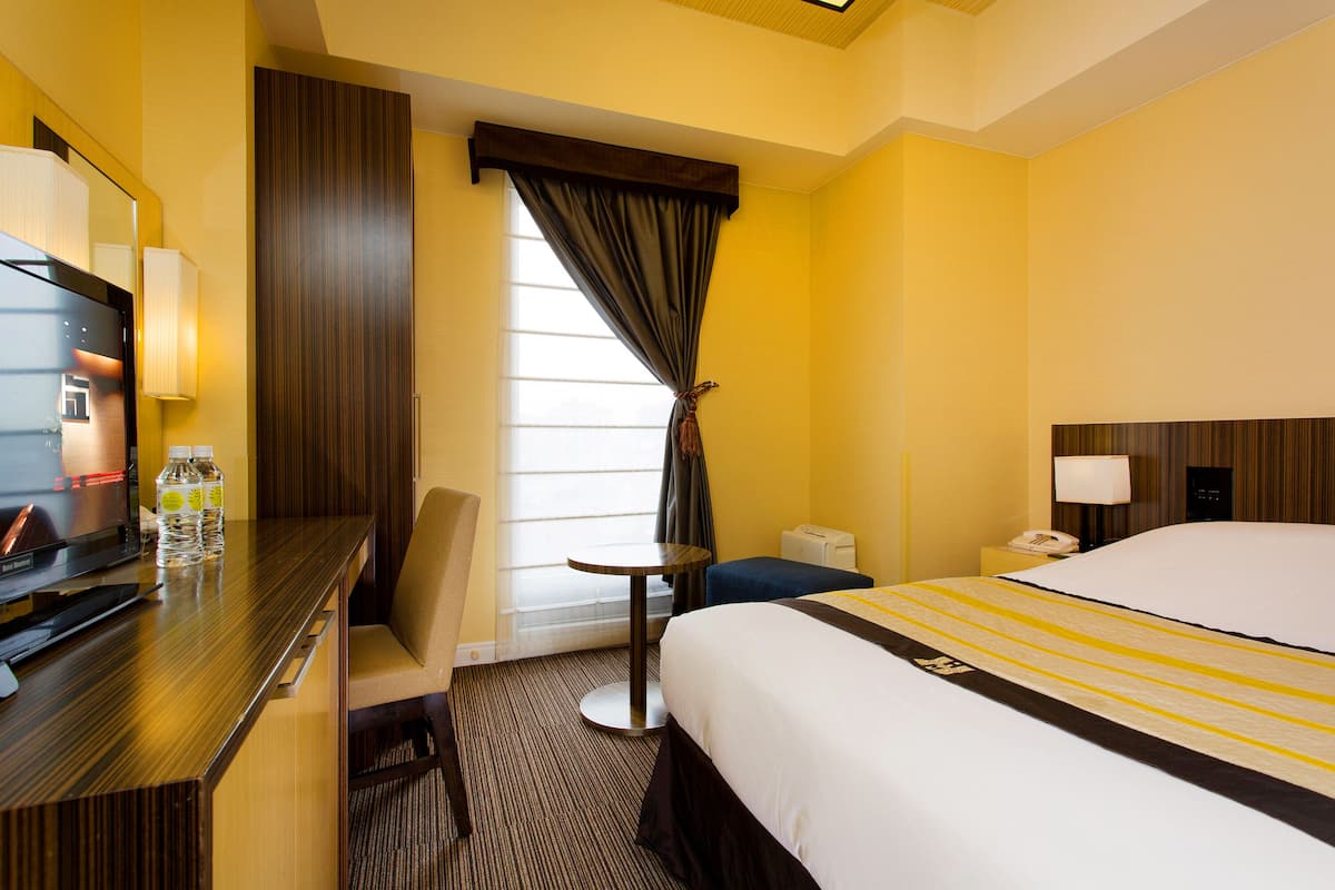A hotel room with a bed, a desk with a computer, a chair, a small table, and a window with curtains.