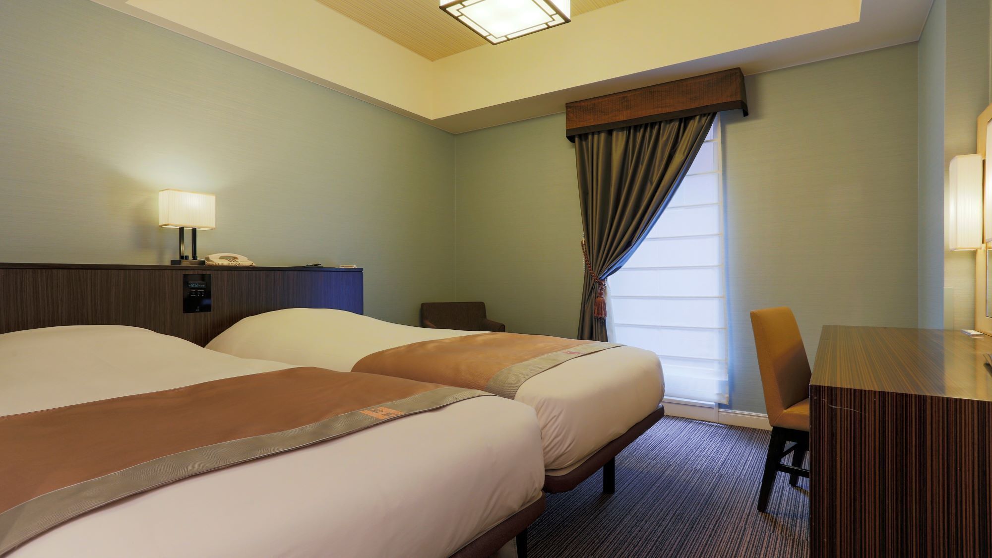 A hotel room with two beds, a desk, a chair, a lamp, and a window with curtains.