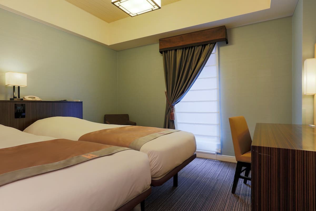 Une chambre d’hôtel avec deux lits, un bureau, une chaise, une lampe et une fenêtre avec des rideaux.