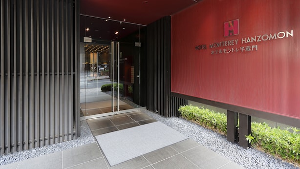 A modern building entrance with a red wall, a glass door, and a metal fence. - Hotel Monterey Hanzomon (Tokyo)