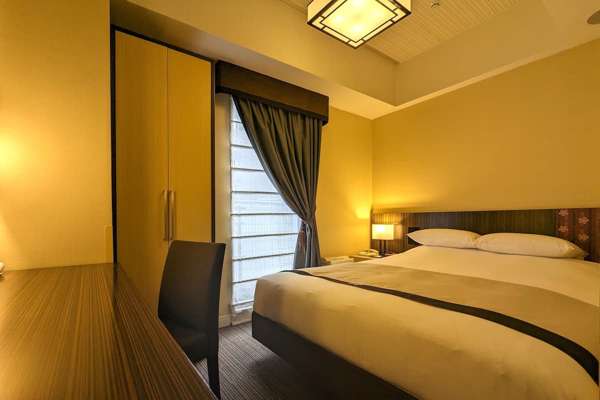 A hotel room with a bed, a desk, a chair, a nightstand, a lamp, and a window with curtains.