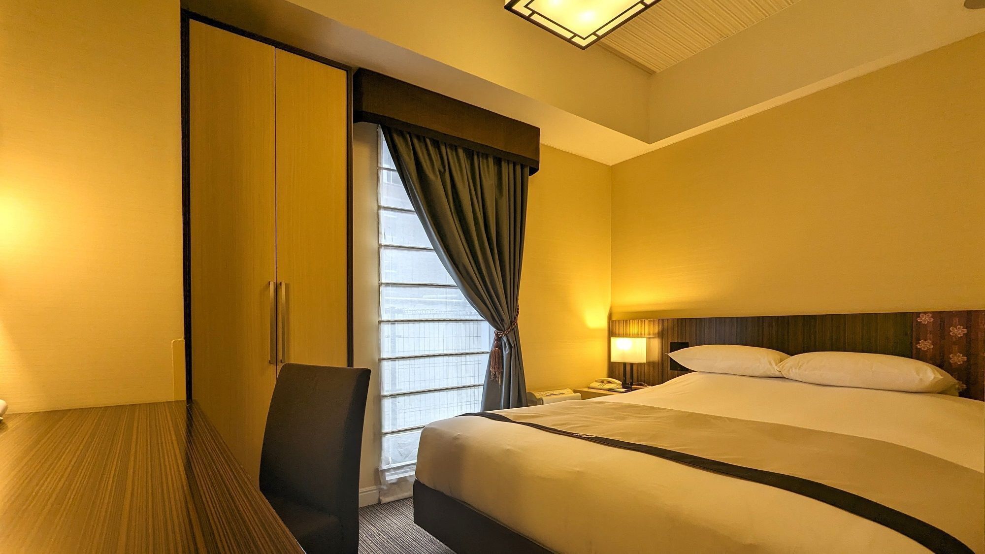 A hotel room with a bed, a desk, a chair, a nightstand, a lamp, and a window with curtains.