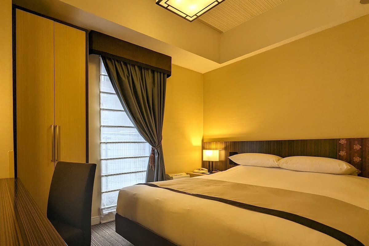 A hotel room with a bed, a desk, a chair, a nightstand, a lamp, and a window with curtains.