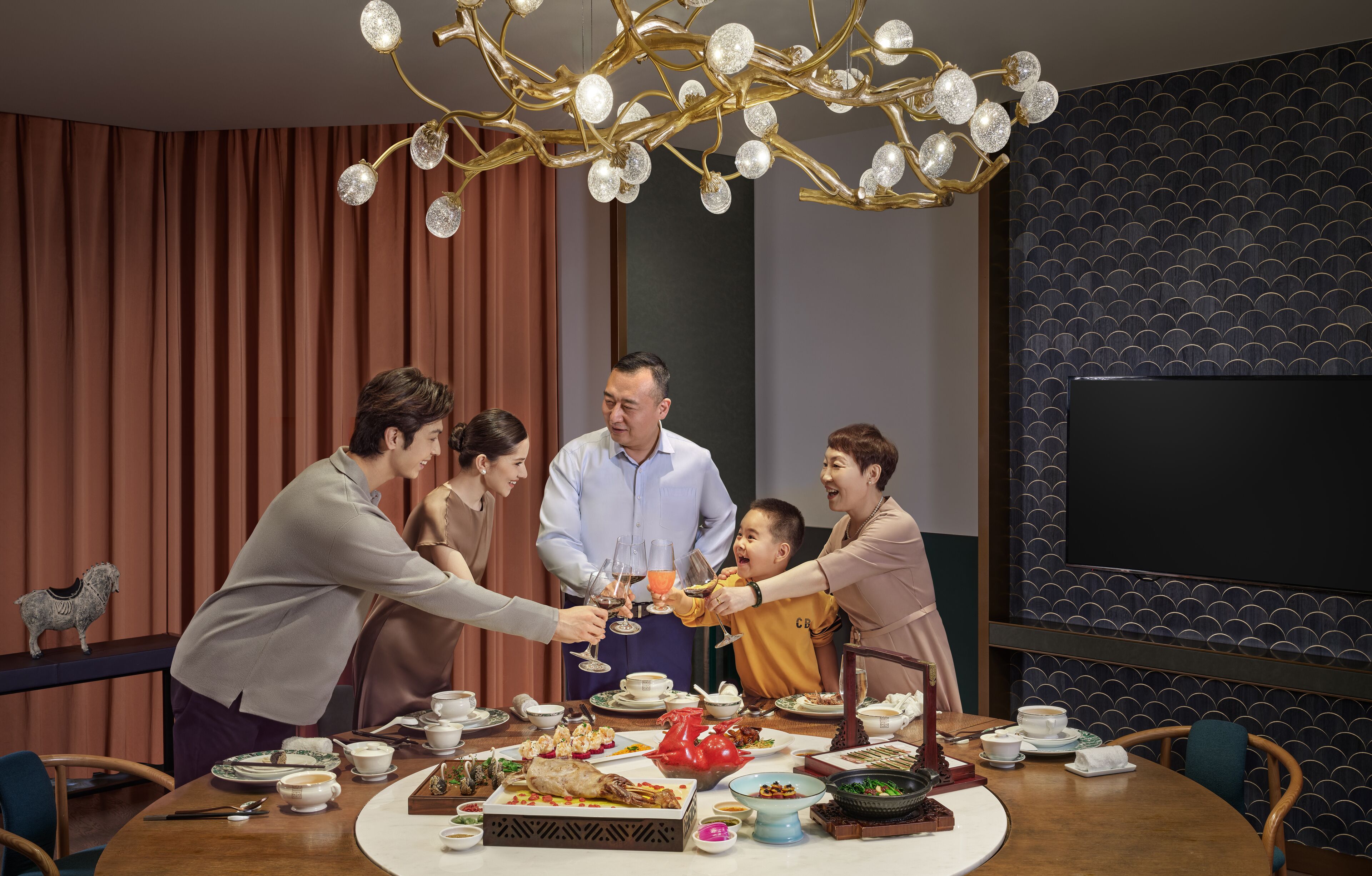 A group of people toasting around a table with various dishes and drinks, in a room with a modern chandelier and patterned wallpaper.